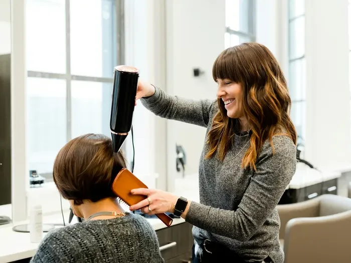 Smiling hair stylist blow dries a client’s short hair with a brush in a bright modern salon, showing local service